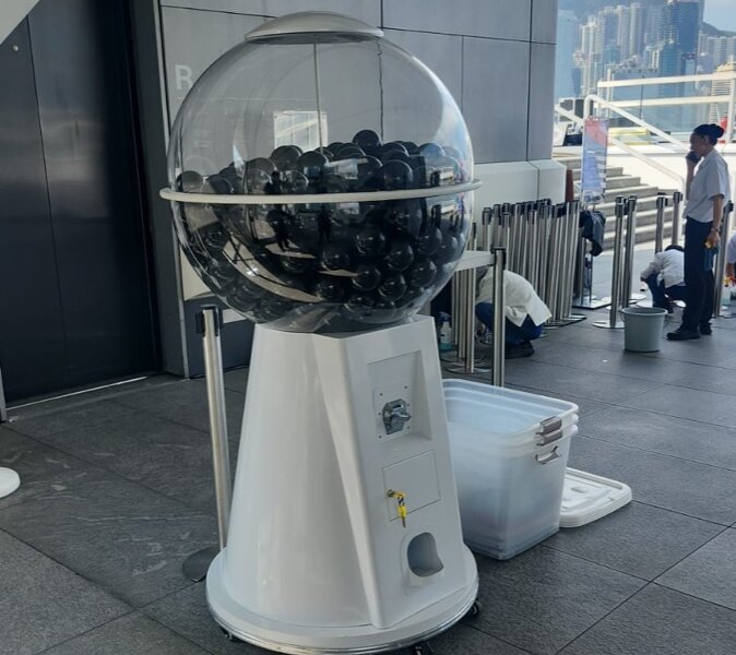 戶外平台的升降機口旁放著一部白色底座的大圓扭蛋機，透明圓球內裝滿黑色扭蛋，底座有轆，四周有伸縮圍欄，遠處可見山景與高樓